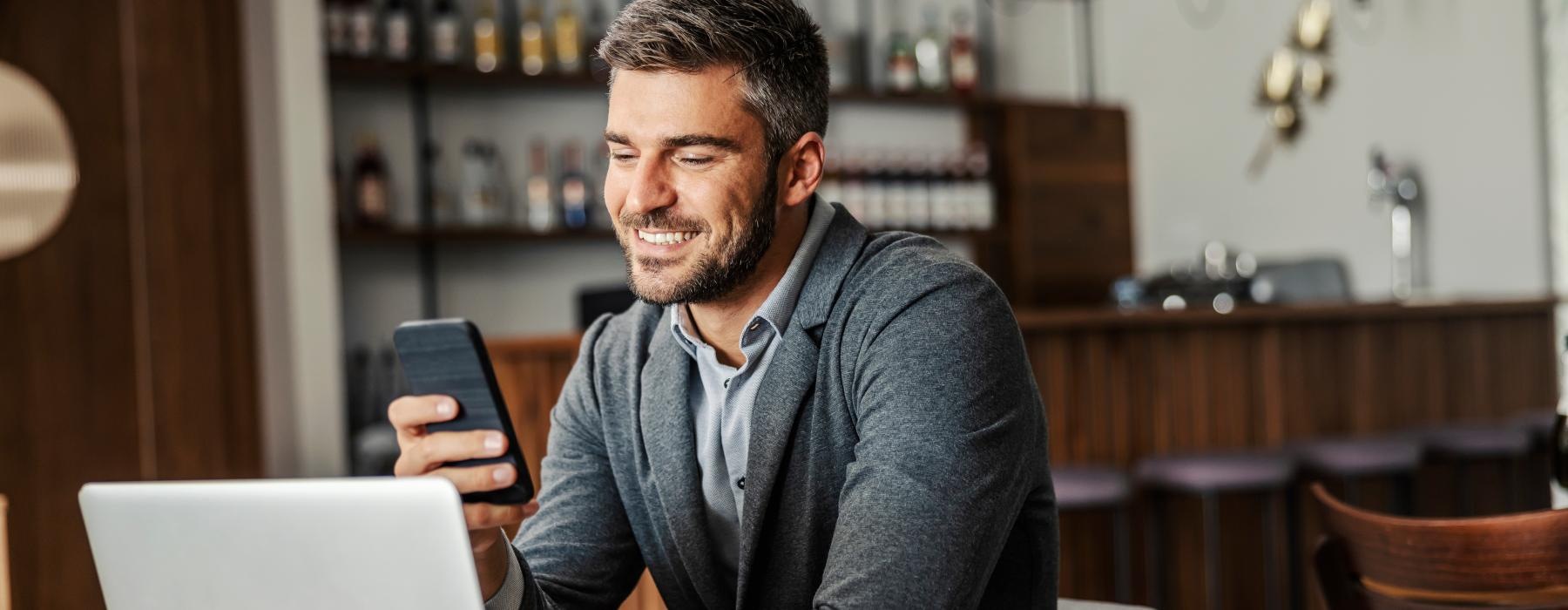 a man looks at his phone while sitting at a table with a laptop and a cup of coffee